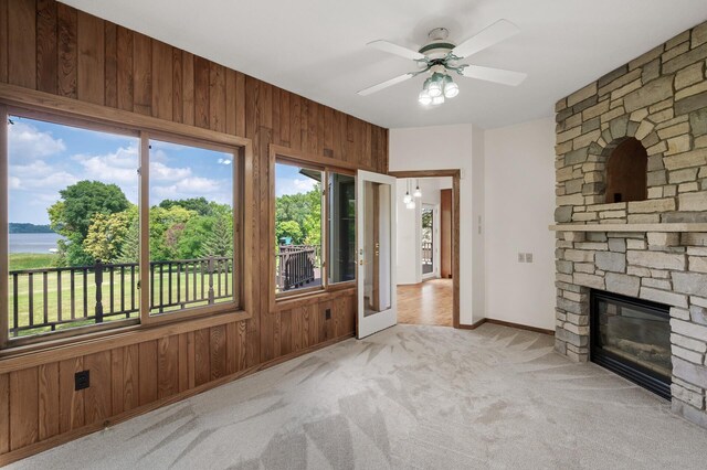 unfurnished living room featuring a fireplace, a water view, wooden walls, ceiling fan, and light colored carpet