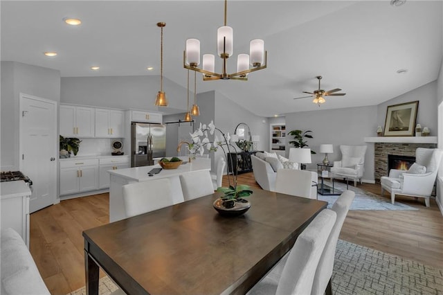 dining area with light wood-type flooring, ceiling fan with notable chandelier, a fireplace, and vaulted ceiling