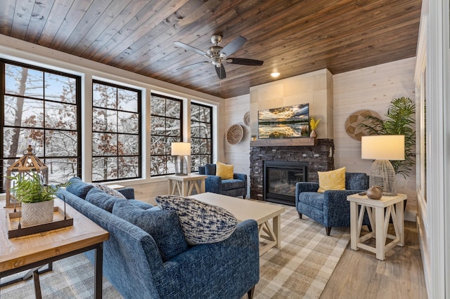 living room with ceiling fan, wooden ceiling, wooden walls, a stone fireplace, and light wood-type flooring