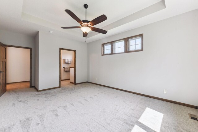 unfurnished bedroom featuring a raised ceiling, ensuite bath, ceiling fan, and light colored carpet