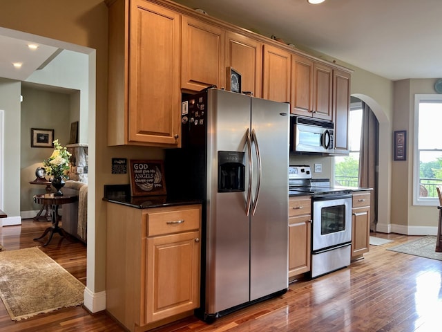 kitchen featuring arched walkways, stainless steel appliances, dark countertops, and wood finished floors