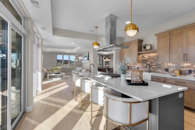 kitchen featuring pendant lighting, island range hood, a kitchen bar, a tray ceiling, and light stone countertops