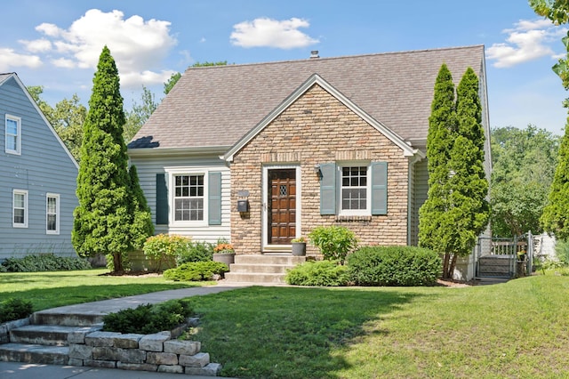 new england style home with a shingled roof, a gate, stone siding, and a front lawn