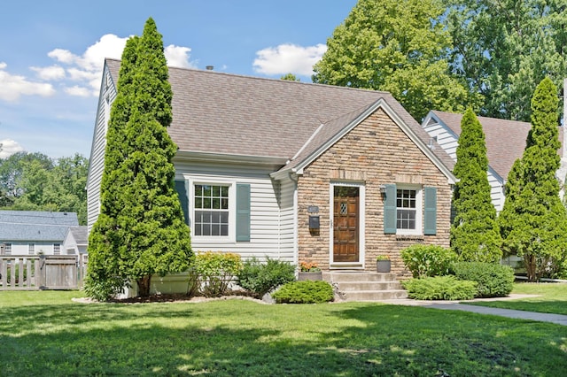 cape cod home featuring stone siding, a front lawn, a shingled roof, and fence