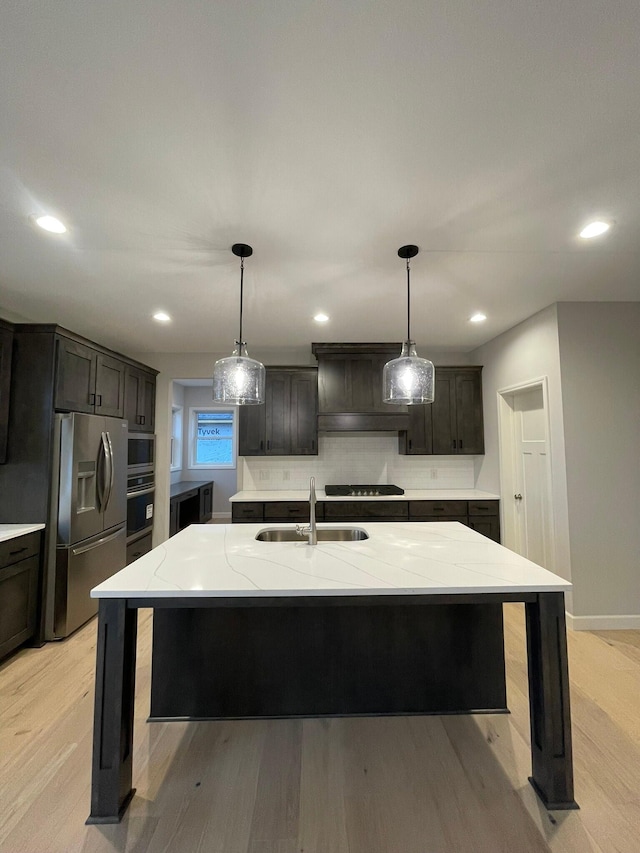 kitchen with sink, an island with sink, backsplash, appliances with stainless steel finishes, and dark brown cabinetry