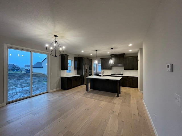 kitchen with an island with sink, light wood-style flooring, a sink, light countertops, and a notable chandelier