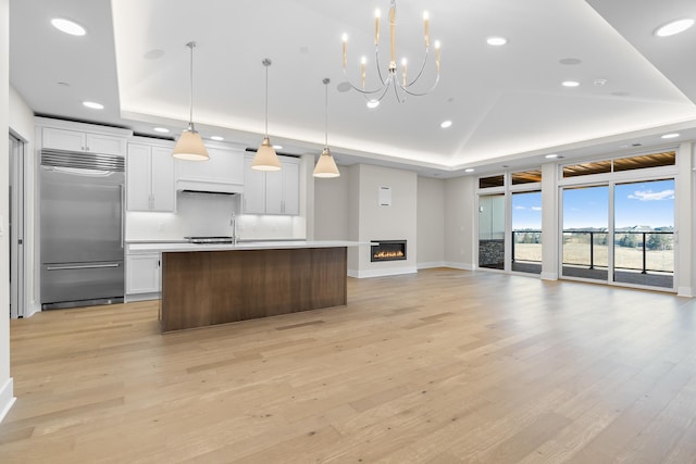 kitchen with stainless steel built in refrigerator, a raised ceiling, pendant lighting, white cabinetry, and an island with sink