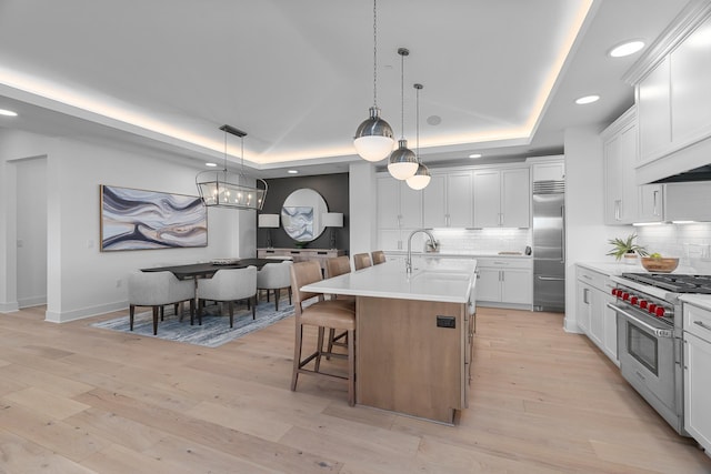 kitchen featuring premium appliances, an island with sink, decorative light fixtures, backsplash, and a raised ceiling