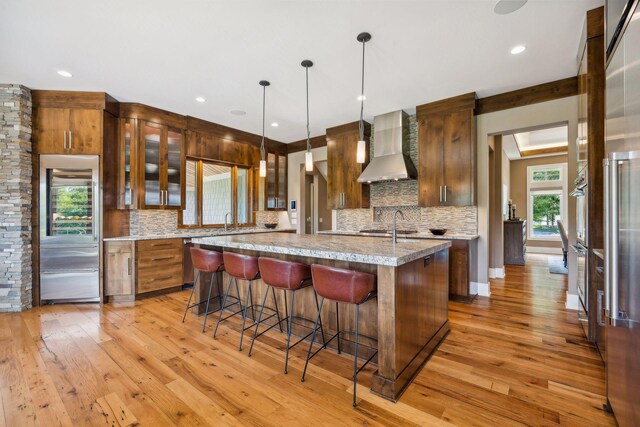 kitchen featuring decorative light fixtures, wall chimney range hood, a center island with sink, light hardwood / wood-style floors, and backsplash