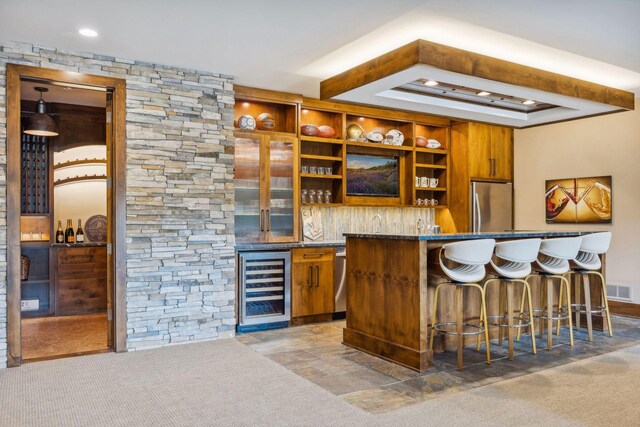 bar featuring carpet flooring, dark stone counters, beverage cooler, stainless steel fridge, and a raised ceiling