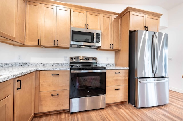 kitchen with appliances with stainless steel finishes, light brown cabinetry, vaulted ceiling, light stone counters, and light hardwood / wood-style flooring