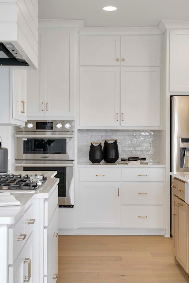 kitchen featuring light hardwood / wood-style flooring, tasteful backsplash, white cabinets, premium range hood, and appliances with stainless steel finishes