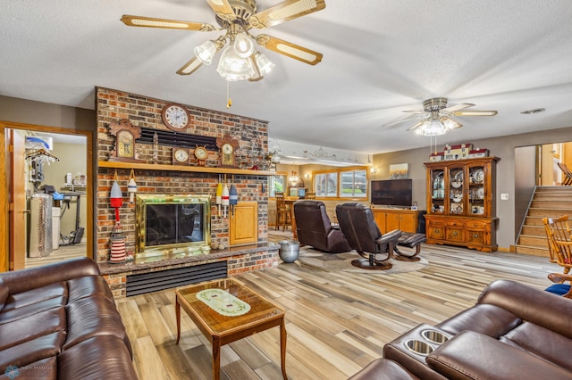 living room with ceiling fan, a textured ceiling, a brick fireplace, and light hardwood / wood-style floors