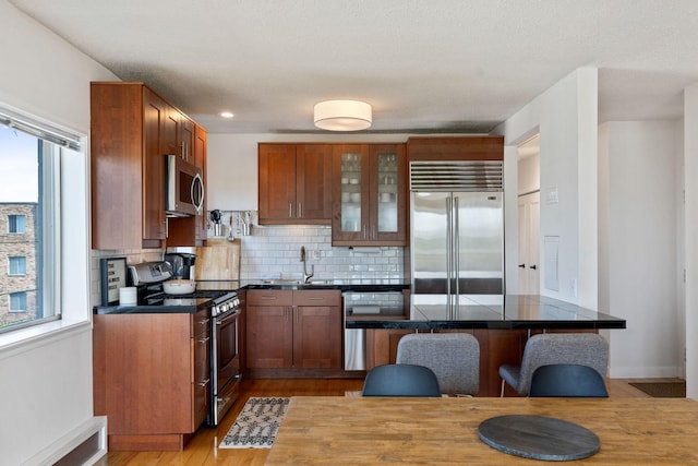 kitchen with dark countertops, glass insert cabinets, appliances with stainless steel finishes, brown cabinets, and a sink