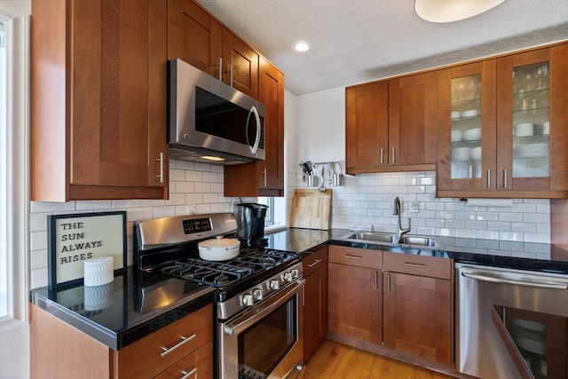 kitchen featuring dark countertops, appliances with stainless steel finishes, brown cabinetry, glass insert cabinets, and a sink