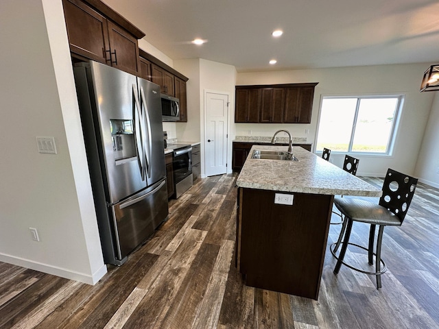 kitchen with dark brown cabinetry, a kitchen island with sink, a sink, stainless steel appliances, and dark wood-style floors