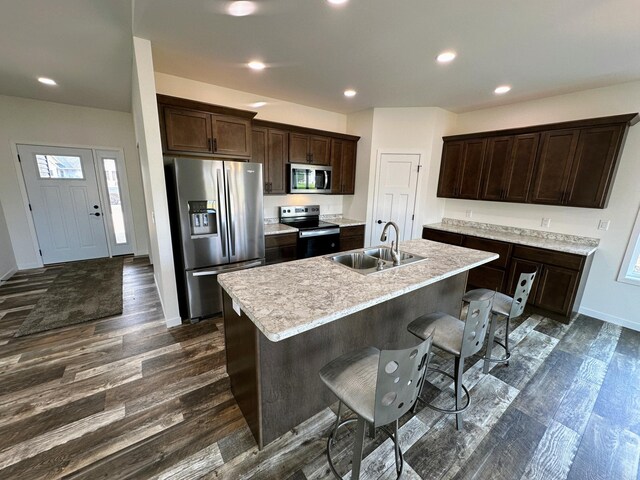 kitchen with dark brown cabinetry, dark wood-style flooring, a sink, stainless steel appliances, and a center island with sink