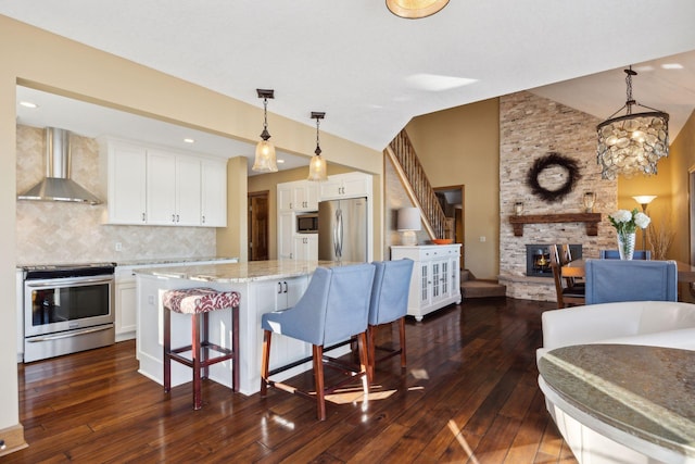 kitchen with a kitchen island, appliances with stainless steel finishes, white cabinets, hanging light fixtures, and wall chimney range hood