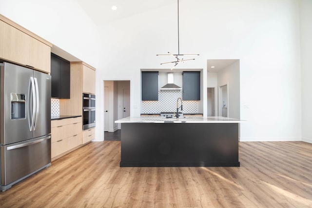 kitchen featuring appliances with stainless steel finishes, a center island with sink, wall chimney range hood, and light wood-type flooring