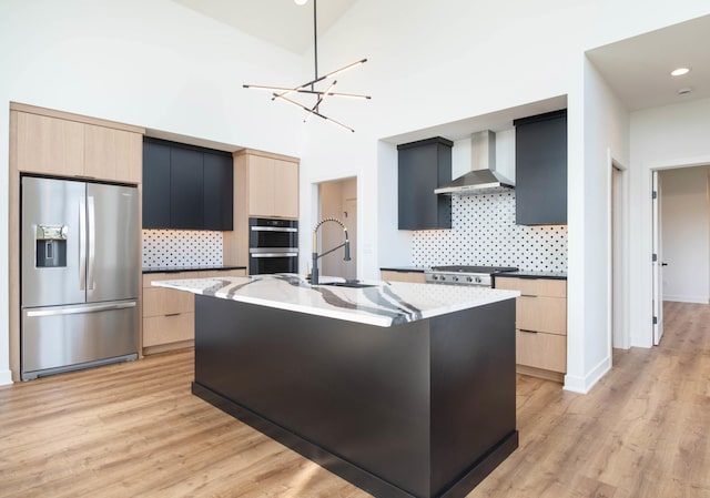 kitchen featuring wall chimney range hood, an island with sink, light stone countertops, light wood-type flooring, and appliances with stainless steel finishes