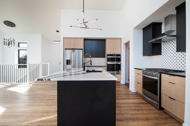 kitchen with a kitchen island with sink, wall chimney range hood, stainless steel appliances, decorative light fixtures, and light brown cabinetry