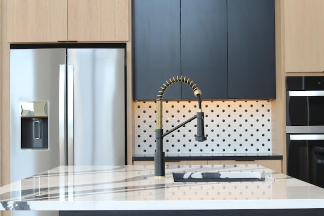 kitchen with light stone countertops, an island with sink, backsplash, stainless steel fridge, and light brown cabinets