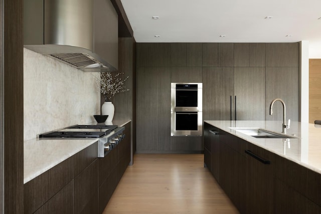 kitchen with wall chimney range hood, sink, light hardwood / wood-style flooring, appliances with stainless steel finishes, and dark brown cabinets