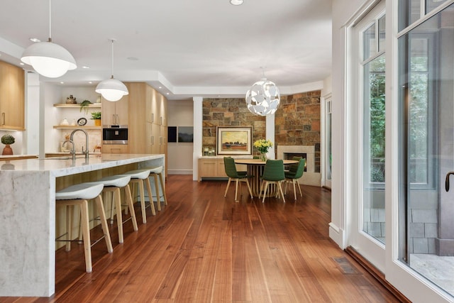 kitchen featuring light brown cabinetry, a stone fireplace, hanging light fixtures, and dark hardwood / wood-style flooring