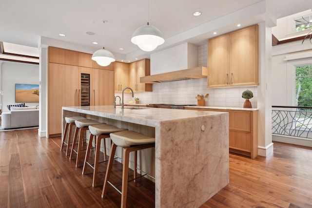 kitchen with hardwood / wood-style flooring, a center island with sink, pendant lighting, light brown cabinetry, and premium range hood