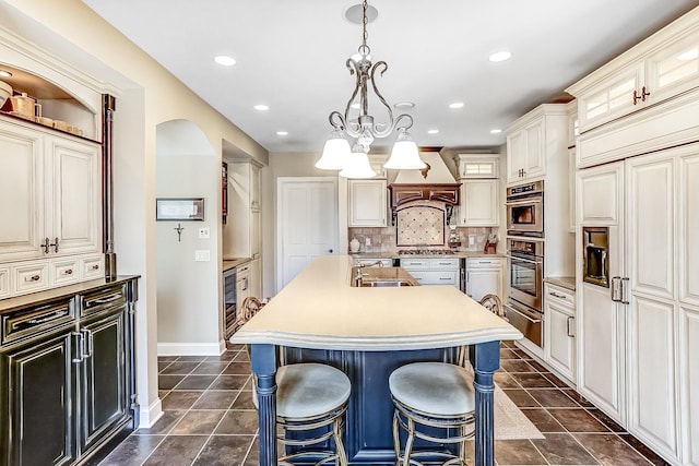 kitchen featuring custom exhaust hood, a center island with sink, backsplash, and dark tile patterned floors