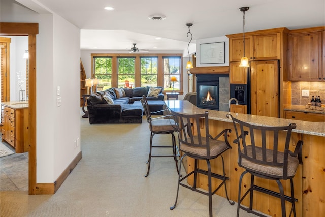 kitchen with light carpet, brown cabinetry, decorative backsplash, a glass covered fireplace, and a kitchen breakfast bar