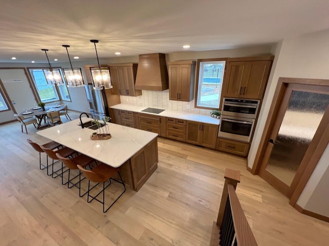 kitchen with a breakfast bar area, appliances with stainless steel finishes, hanging light fixtures, custom range hood, and a center island with sink