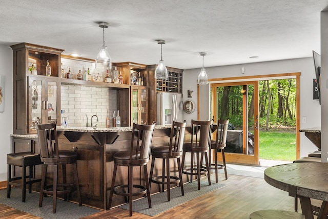 bar with pendant lighting, stainless steel refrigerator with ice dispenser, a textured ceiling, wood finished floors, and indoor wet bar