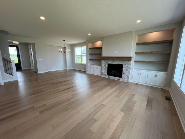 unfurnished living room with built in shelves, a stone fireplace, a chandelier, light hardwood / wood-style flooring, and a textured ceiling