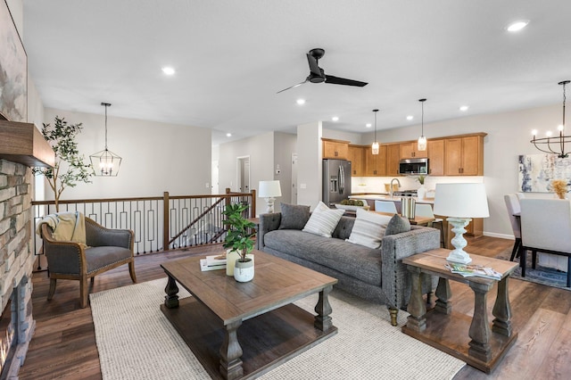 living room with dark wood-type flooring, ceiling fan with notable chandelier, a stone fireplace, and sink