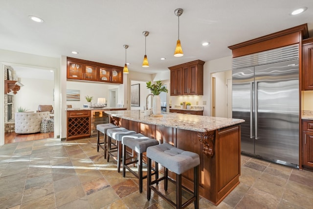 kitchen with a breakfast bar area, a kitchen island with sink, hanging light fixtures, light stone countertops, and built in fridge