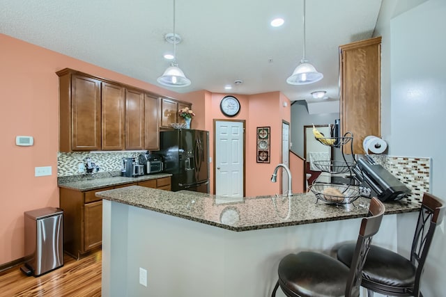 kitchen featuring a peninsula, black fridge, light wood-type flooring, decorative backsplash, and dark stone counters