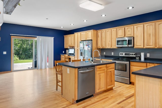 kitchen with a sink, a center island with sink, stainless steel appliances, light brown cabinets, and dark countertops