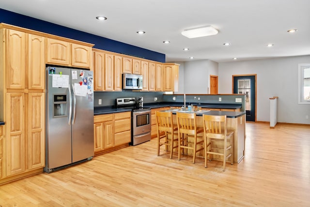 kitchen with a kitchen breakfast bar, light brown cabinetry, dark countertops, light wood-style flooring, and appliances with stainless steel finishes