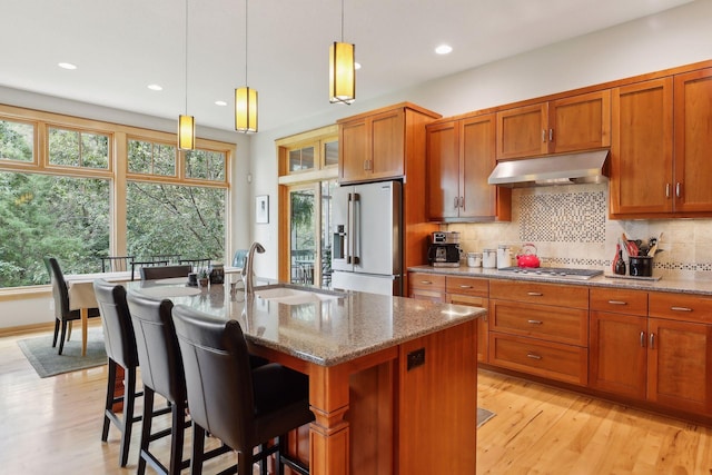 kitchen with light stone counters, a sink, stainless steel appliances, under cabinet range hood, and backsplash