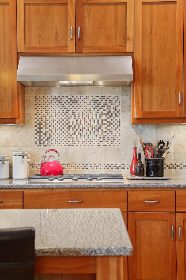 kitchen featuring wall chimney range hood, stainless steel gas cooktop, light stone counters, decorative backsplash, and brown cabinets