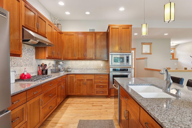 kitchen with under cabinet range hood, a sink, appliances with stainless steel finishes, brown cabinetry, and light wood finished floors