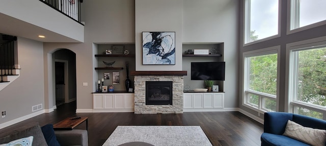 living room featuring built in shelves, a towering ceiling, a stone fireplace, and dark wood-type flooring