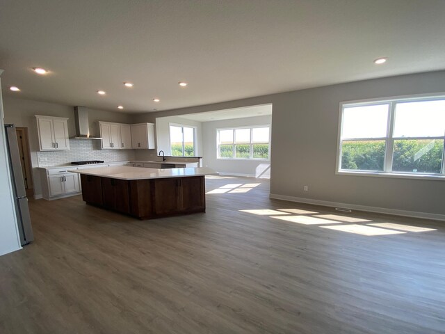 kitchen featuring wall chimney range hood, wood-type flooring, white cabinets, and a center island with sink