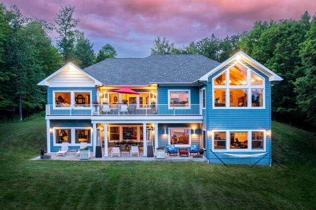 back house at dusk featuring a balcony, a patio, outdoor lounge area, and a lawn