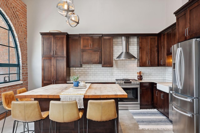 kitchen with wall chimney exhaust hood, butcher block countertops, appliances with stainless steel finishes, a kitchen island, and decorative backsplash