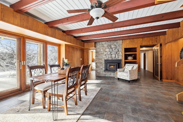 dining area with beamed ceiling, wooden walls, a stone fireplace, and ceiling fan