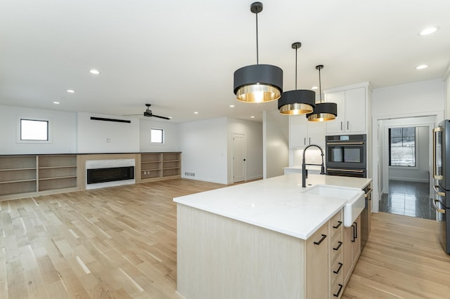 kitchen featuring decorative light fixtures, light wood-type flooring, an island with sink, black oven, and white cabinets