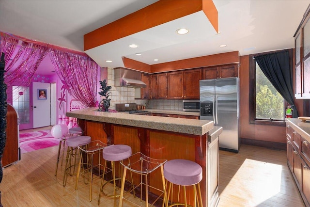 kitchen with tasteful backsplash, stainless steel appliances, light hardwood / wood-style floors, wall chimney range hood, and a kitchen bar