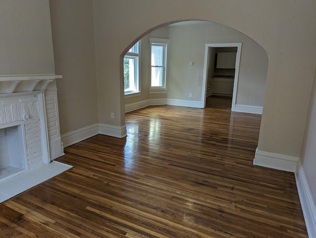 unfurnished living room with baseboards, arched walkways, a brick fireplace, and dark wood-style flooring